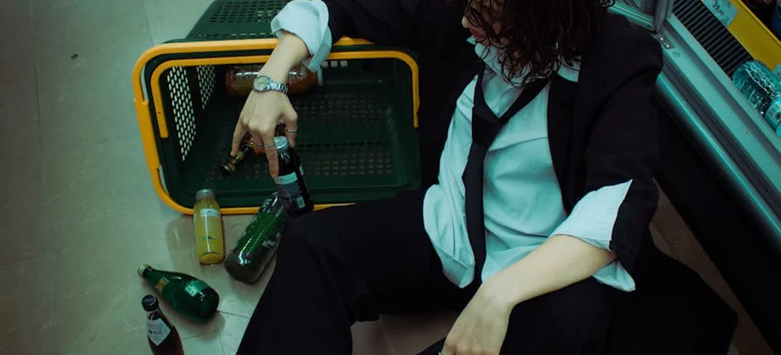 Person wearing a black suit with a white shirt and a loosely tied dark tie, seated on the floor with bottles nearby.