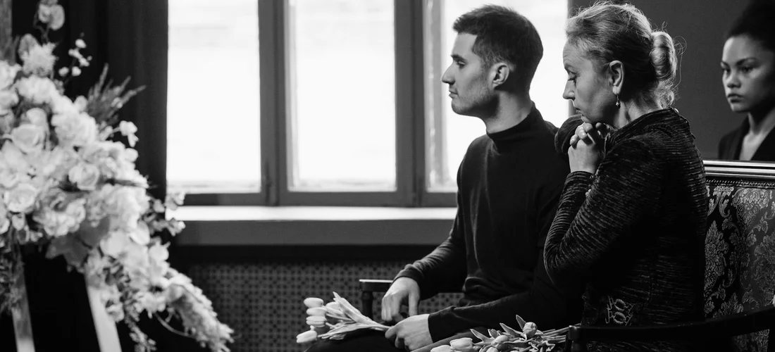 Three mourners sit solemnly in a funeral setting with flowers nearby, reflecting on the emotional weight and personal limits of attending.
