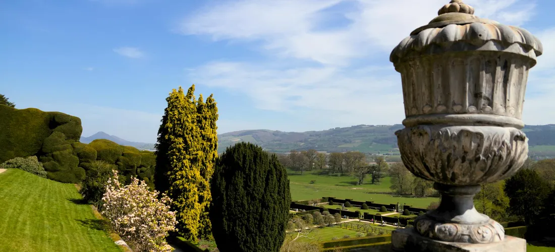 Ornate stone garden urn on a pedestal overlooking a manicured lawn, trees, and a bright blue sky.