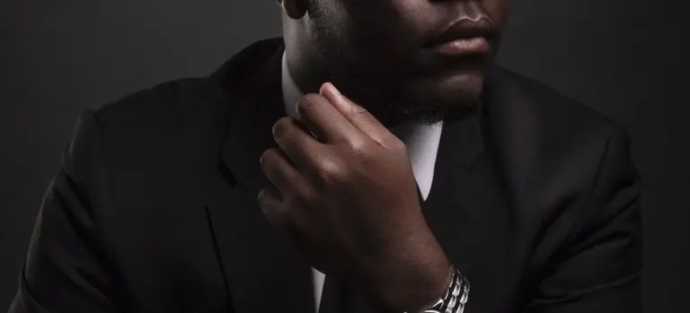 Close-up of a person in a dark suit and white shirt, with a hand resting on the chin.