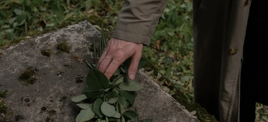A hand resting on a gravestone with a small bouquet of green leaves at a cemetery.
