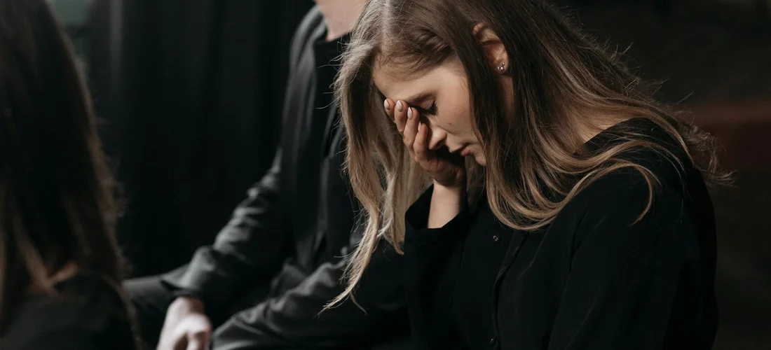 A woman dressed in black sits with her head bowed and hand covering her face at a funeral.