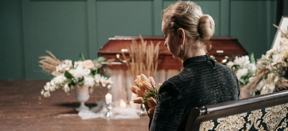A grieving woman in a dark outfit sits on a bench inside a funeral setting, holding a bouquet while facing a casket surrounded by white floral arrangements.