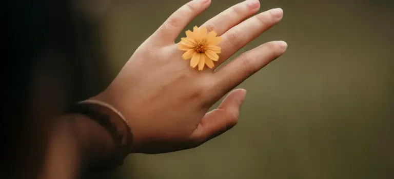 Close-up of a hand with a small yellow-orange flower ring on the middle finger, softly blurred background