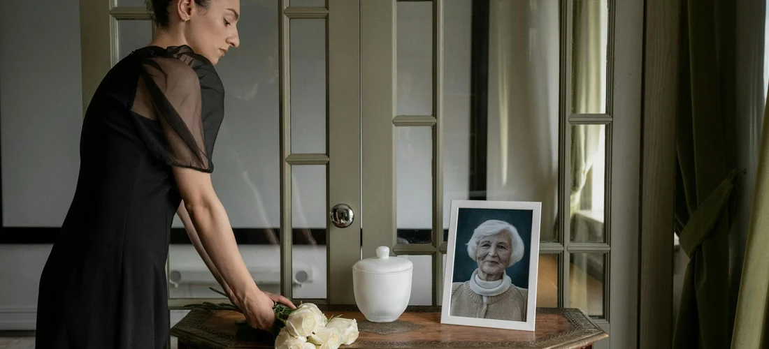A woman in a black dress stands by a wooden table with a framed portrait of an elderly woman, a white vase, and soft light in a quiet room, suggesting memory and tribute.
