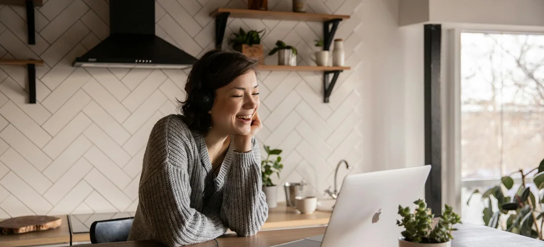 Smiling woman in a gray sweater sits at a kitchen counter with a laptop, preparing to livestream a funeral service from home