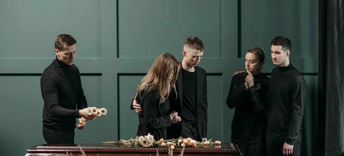 Mourners dressed in black stand around a coffin with floral arrangements, preparing and reflecting on a tribute as part of a funeral service.