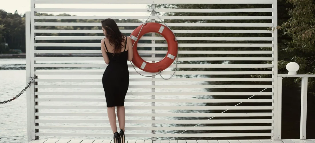 A woman wearing a black knee-length dress stands with her back to the camera on a dock, facing a white slatted barrier with a red lifebuoy.