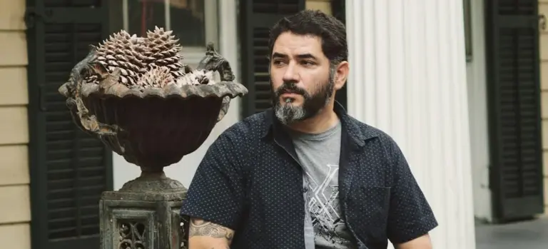 A man with a beard wearing a dark short-sleeve shirt stands outdoors beside a large decorative planter, looking to the side.