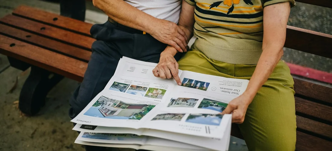 Two people sitting on a wooden park bench and looking at a spread of photos in a newspaper.