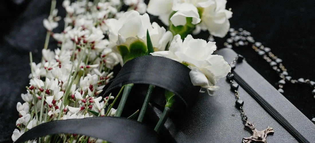 Close-up of a black mortarboard cap adorned with white flowers and a rosary, symbolizing a funeral.