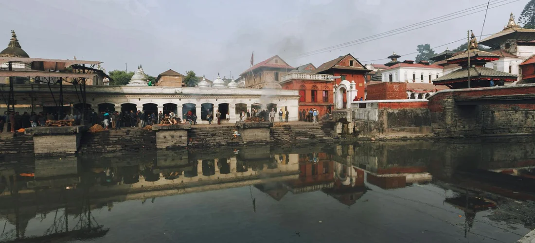 Riverside cremation ghats with temple architecture and groups of people gathered along the water's edge.