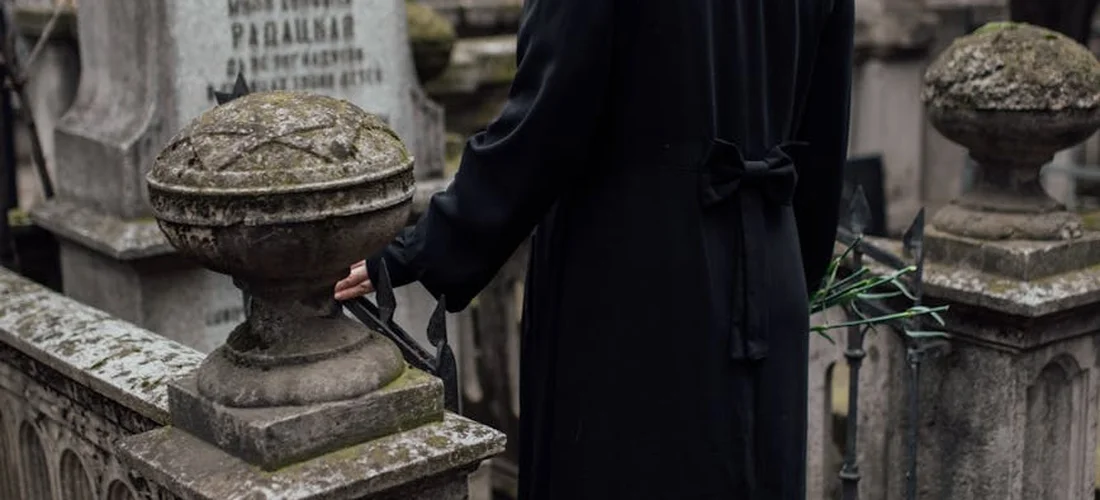 A person in dark clothing stands beside weathered gravestones in a cemetery, reflecting in a quiet, somber scene.
