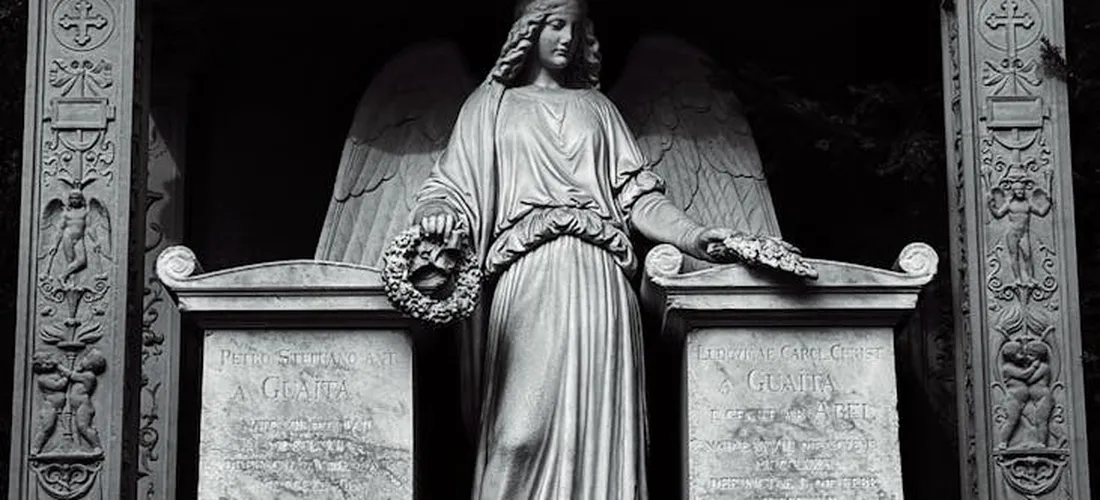 Grayscale photograph of a mourning female statue between two stone tombs in a cemetery.