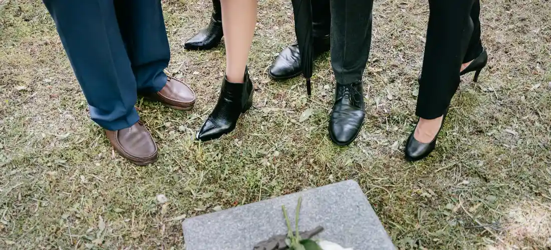 A group of people stands around a grave marker in a grassy cemetery; a small plant rests on the stone.