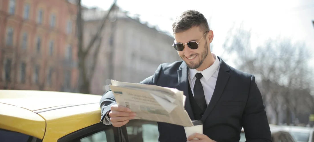 A man in a suit wearing sunglasses reads a newspaper beside a yellow taxi.