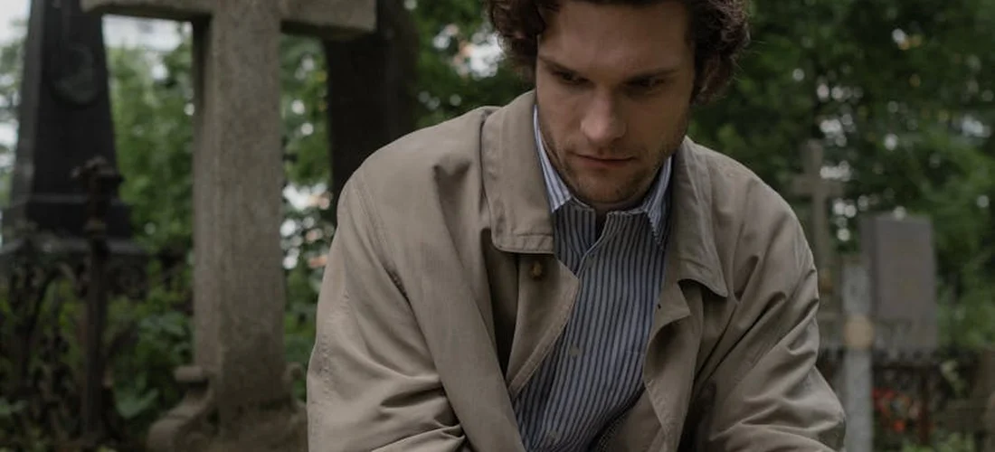 Grieving man sitting in a cemetery, looking down with a somber expression during a funeral setting.
