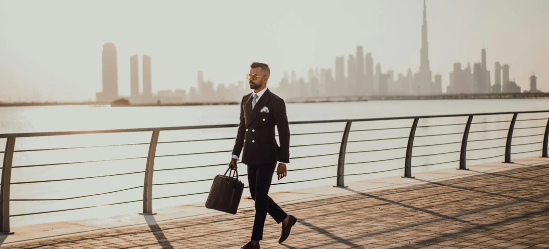 A man in a tailored dark suit walks along a waterfront promenade carrying a briefcase, with a modern city skyline in the distance.