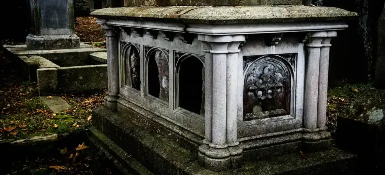 Stone mausoleum with ornate panels in a cemetery, surrounded by fallen leaves.
