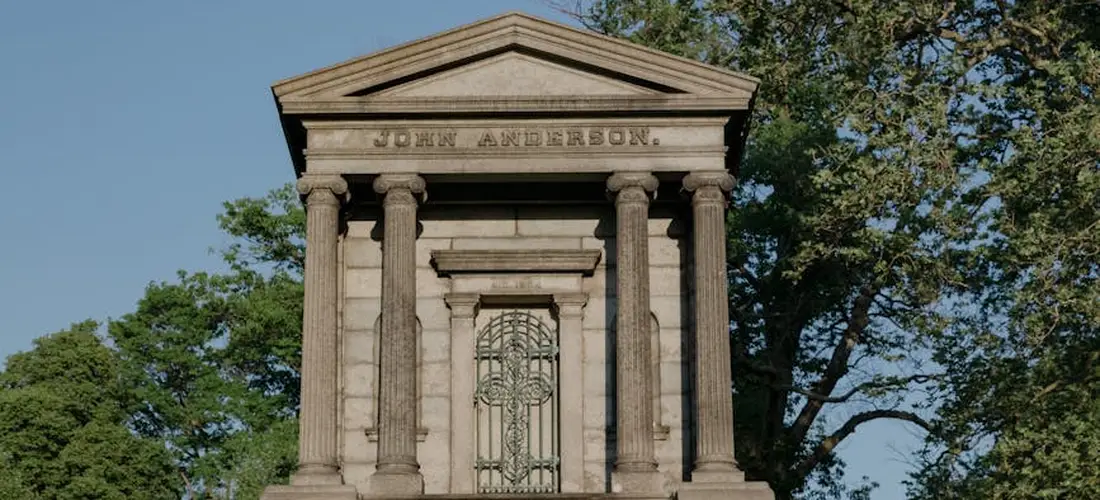 Stone mausoleum with tall columns and a decorative iron doorway surrounded by trees.