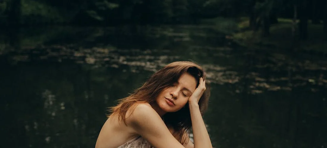 A woman sits by a calm lake, resting her head on her hand, looking contemplative.