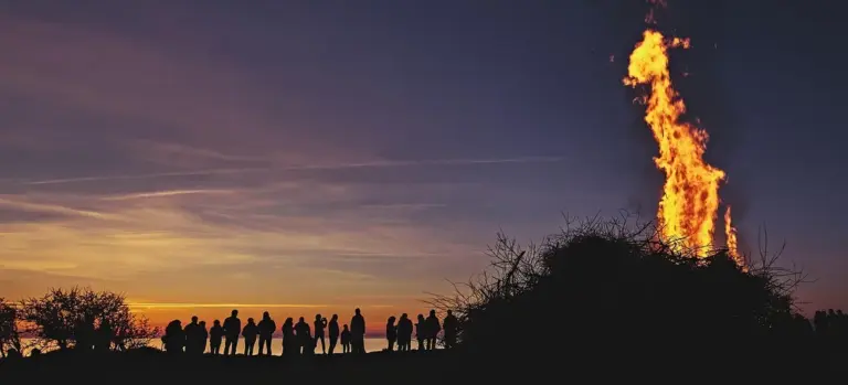 Silhouetted crowd gathered around a tall bonfire on a hill at dusk, with a colorful sky