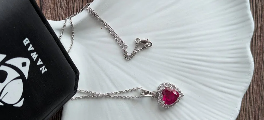 Silver necklace with a red heart-shaped pendant laid on a white plate next to a black presentation box.