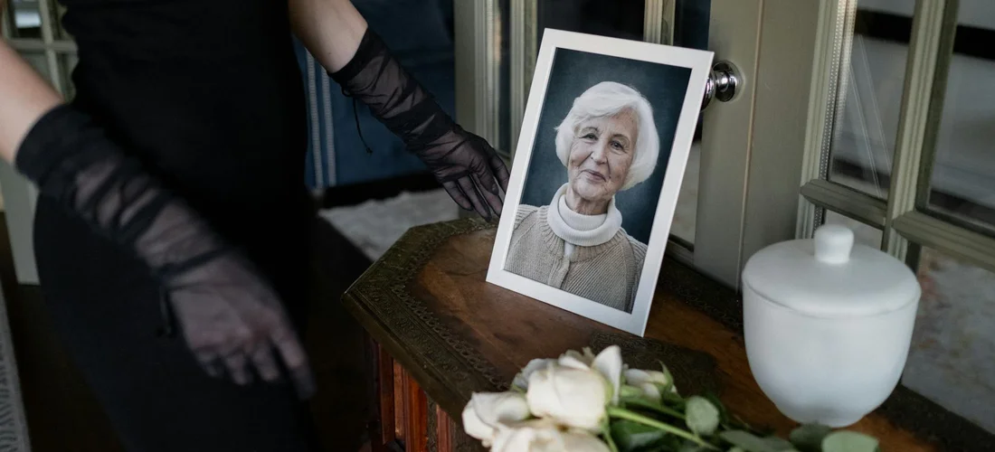 Framed portrait of an elderly woman on a wooden table with white flowers and a white keepsake urn nearby, suggesting a funeral or memorial setup.