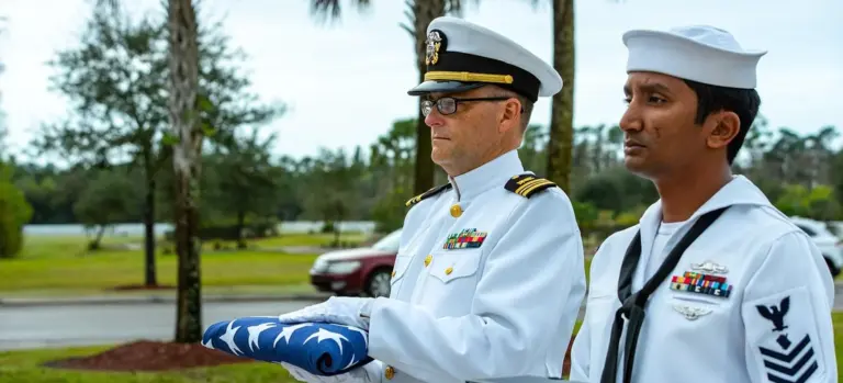 Two uniformed service members standing outdoors, one in a white dress uniform holding a folded flag.