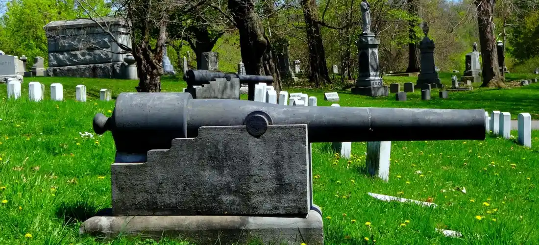 A ceremonial cannon in the foreground at a cemetery, with green grass and rows of white headstones in the background.