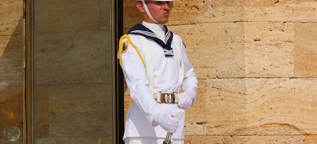 A military honor guard in white dress uniform stands at attention near a doorway, holding a ceremonial sword.