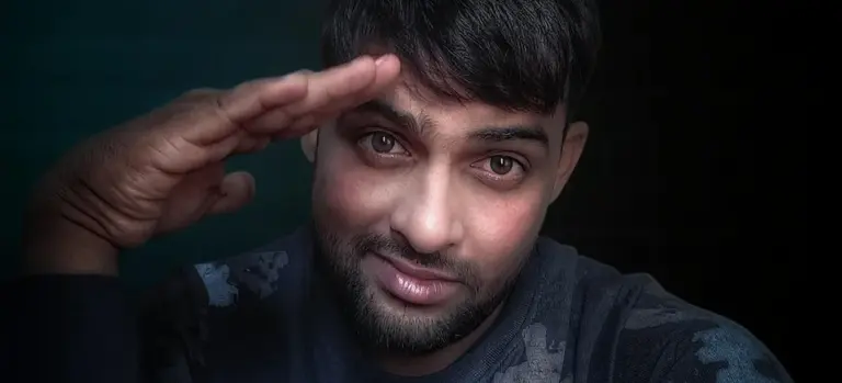 Close-up portrait of a man in casual clothing giving a ceremonial salute with his right hand raised to his temple, dark background.