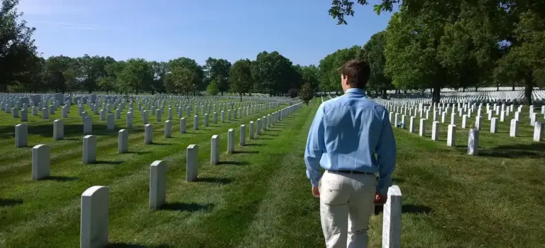 Man walking through rows of white headstones in a green military cemetery, symbolizing veteran burials and related benefits.