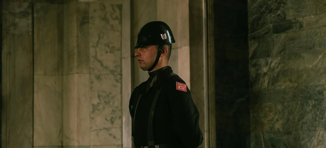Ceremonial military guard at attention in a marble-walled interior, wearing a helmet and dark uniform.