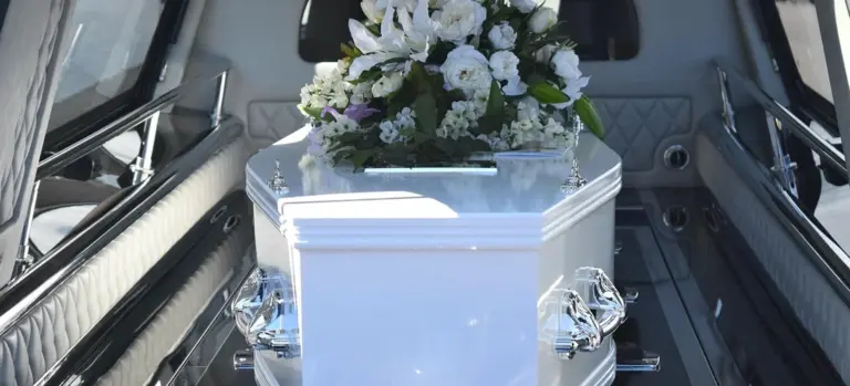 White coffin adorned with flowers inside an open hearse, conveying a solemn funeral setting.