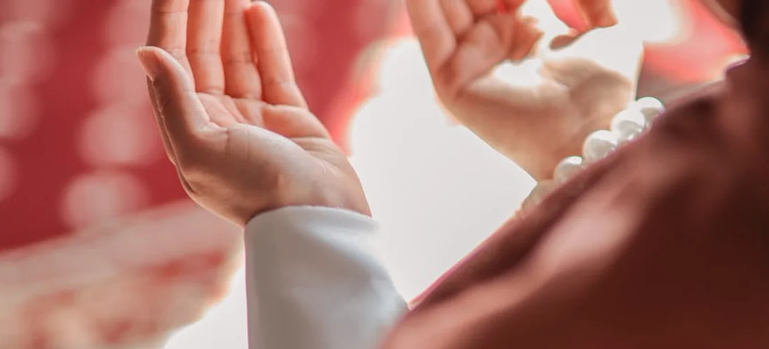 Close-up of hands raised in prayer during a Muslim funeral ritual, with a string of prayer beads visible on the wrist.
