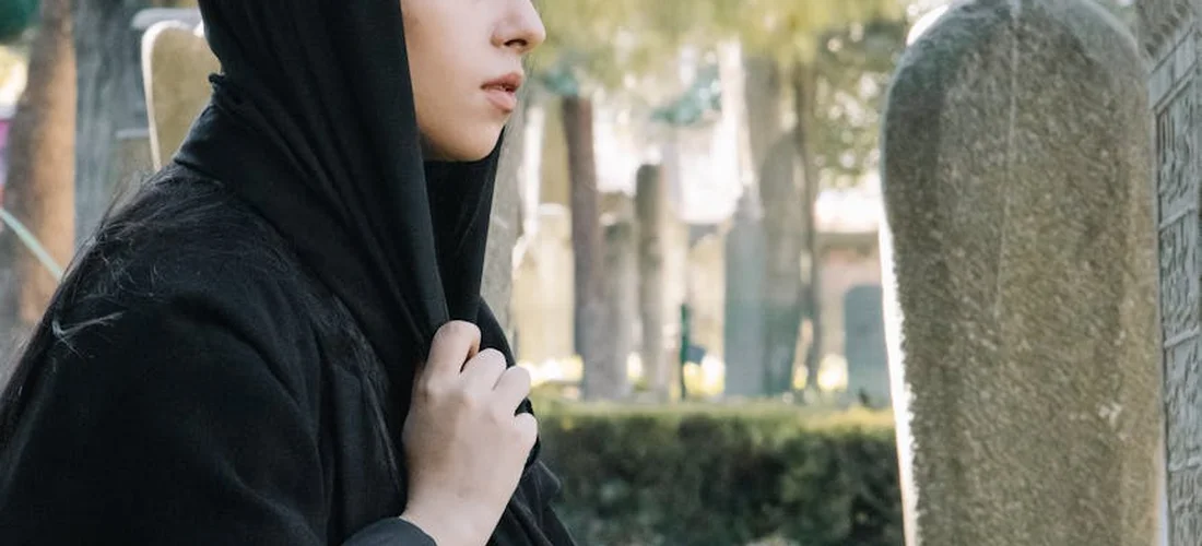 A Muslim woman wearing a black hijab stands beside a gravestone in a cemetery, reflecting near the graves.