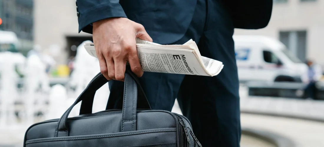 Person in business attire holding a newspaper and a briefcase on a city street