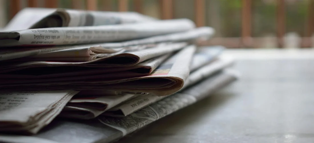 A stack of folded newspapers on a table, representing research and archival sources for obituary writing.