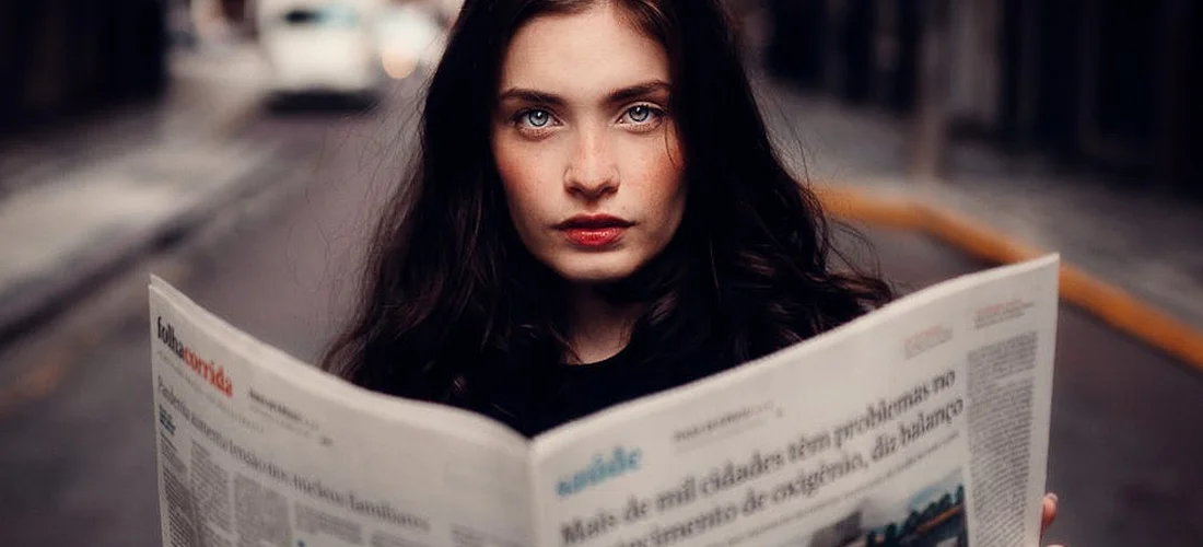 Woman with dark hair reading a newspaper on a city street, focused and pensive.