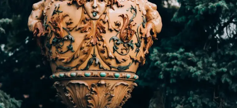 Close-up of an ornate urn with carved floral vines and turquoise accents, set against a dark green, leafy background