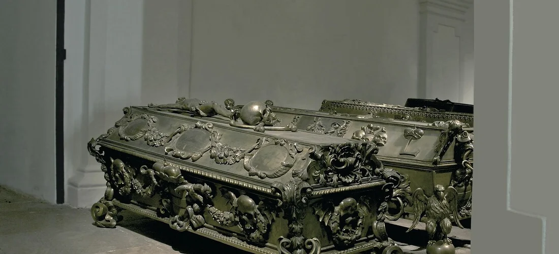 Closed ornate casket resting on the floor in a neutral, well-lit room.
