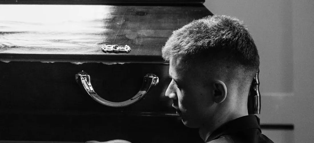 A pallbearer stands beside a closed casket in a somber, black-and-white photograph.