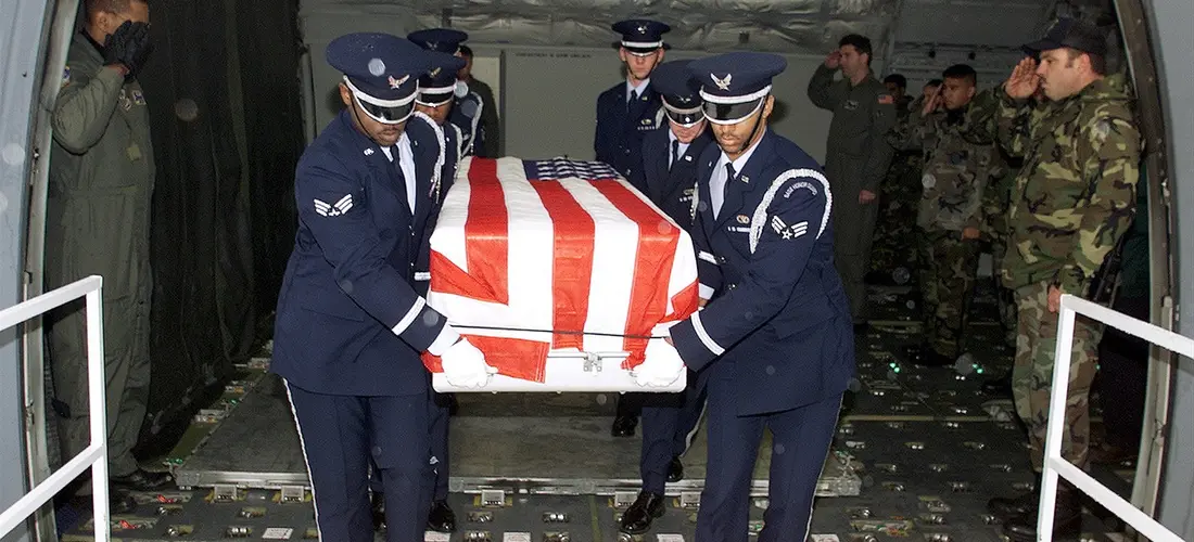 Military pallbearers in dress uniforms carry a flag-draped casket onto a transport, illustrating ceremonial duties.