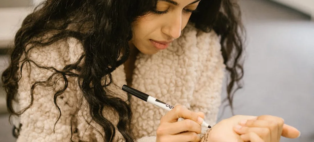Close-up of a person with long dark curly hair wearing a beige fleece jacket, holding a marker and writing on a surface, suggesting note-taking during archival research.