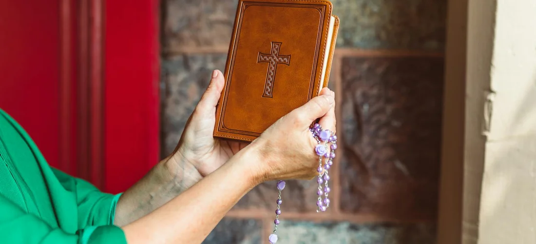 Hands holding a brown prayer book with a cross on the cover and a rosary draped over the book
