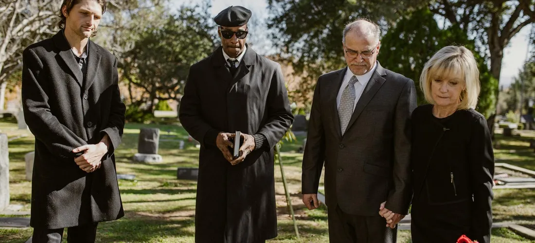 Four attendees in a cemetery, two men in dark attire, a clergyman, and a woman, standing solemnly.