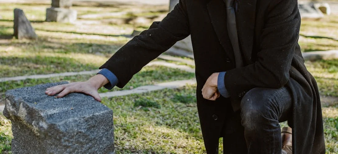 A person in dark clothing kneels beside a gravestone in a cemetery, placing a hand on the stone.