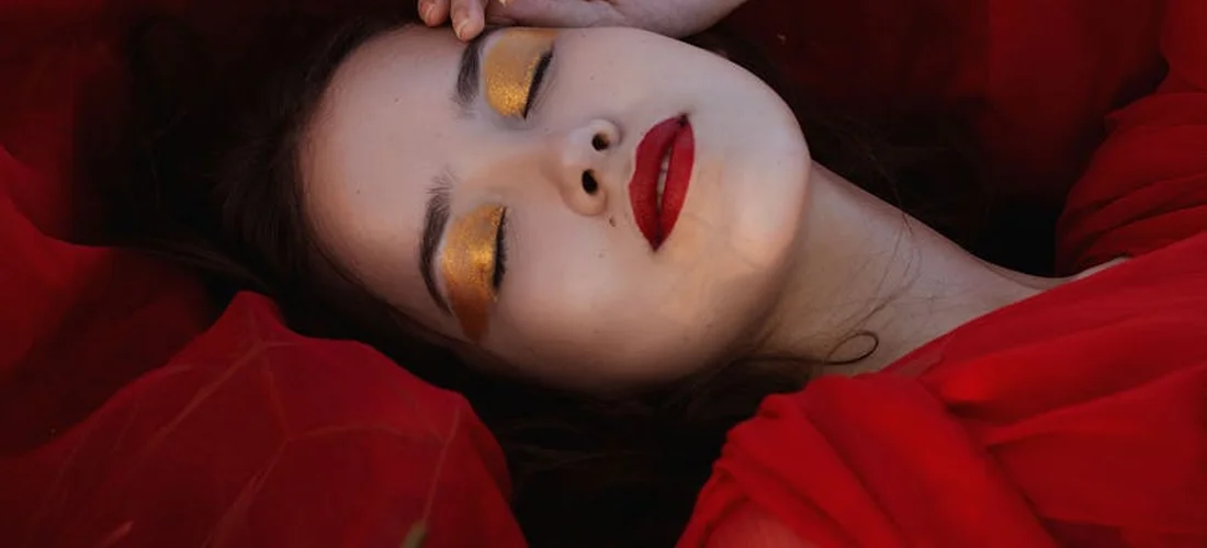 Close-up portrait of a person with red fabric draped around them, wearing gold eyeshadow and bold red lipstick, eyes closed.