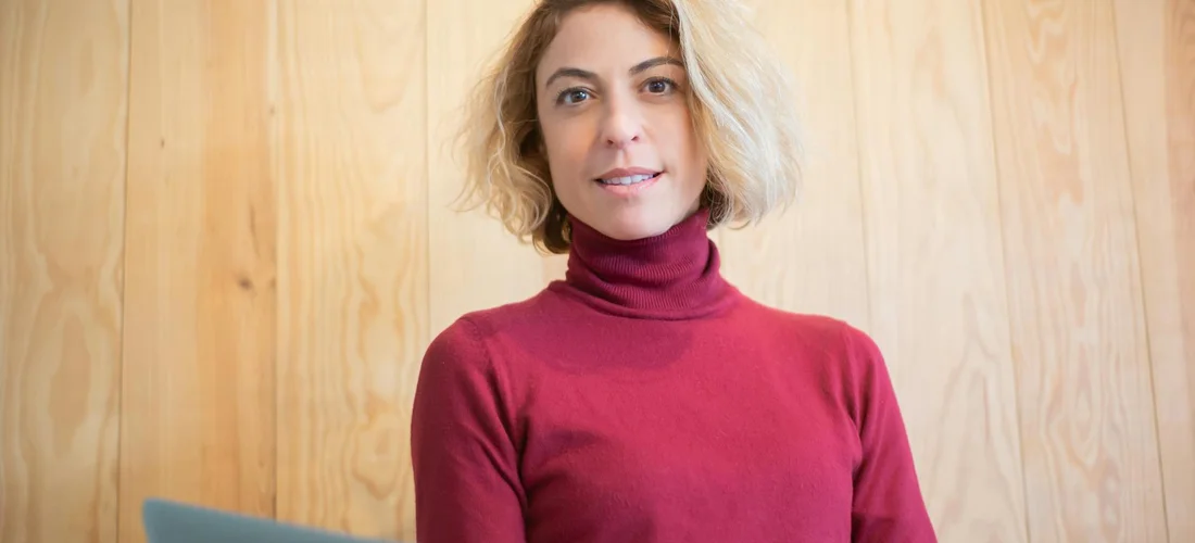 Woman with short blonde hair wearing a burgundy turtleneck, standing against a light wooden wall.